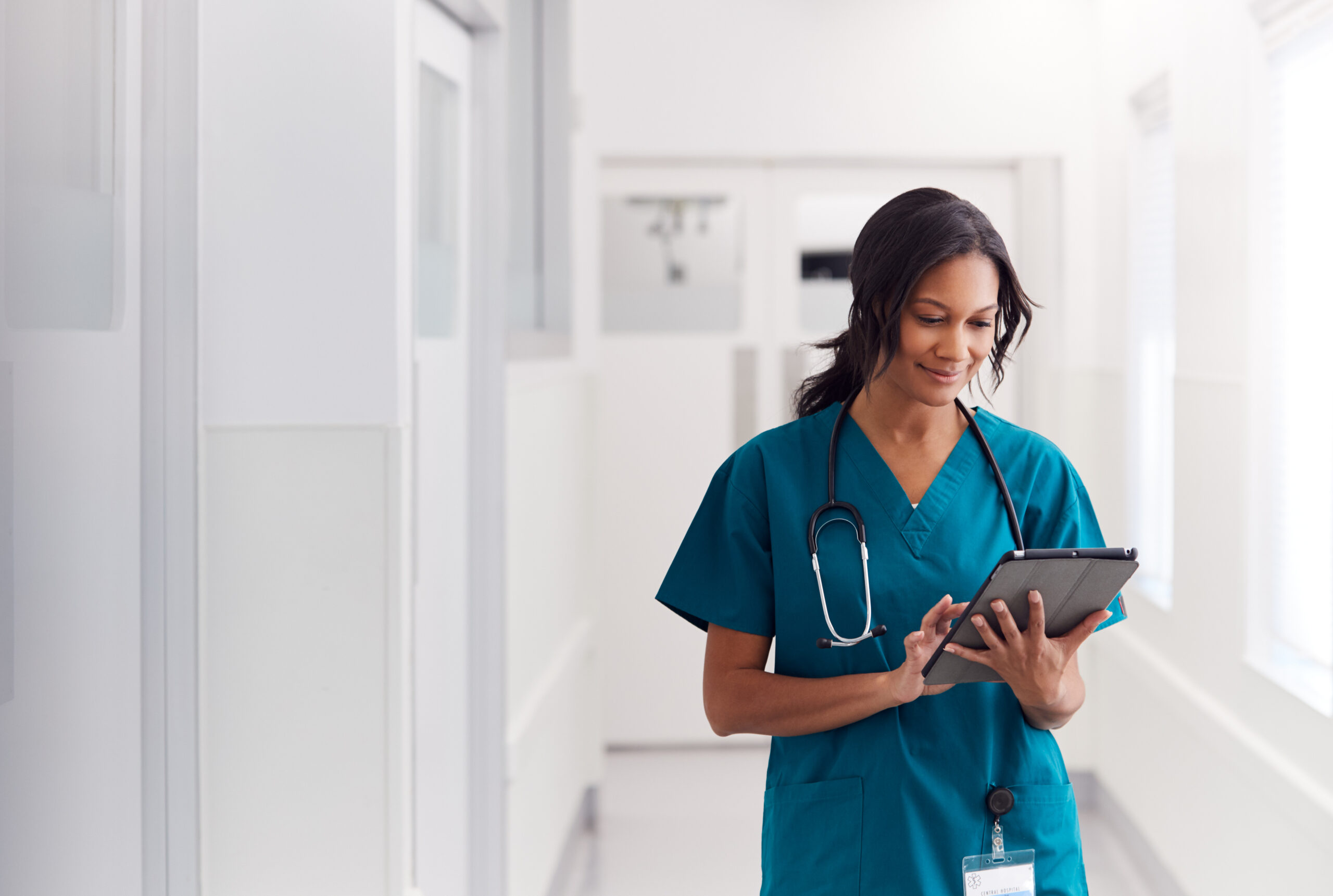 Female Doctor Wearing Scrubs In Hospital Corridor Using Digital Tablet