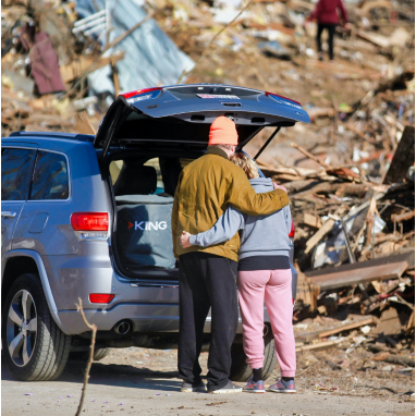 man and woman embracing at back of car in front of rubble